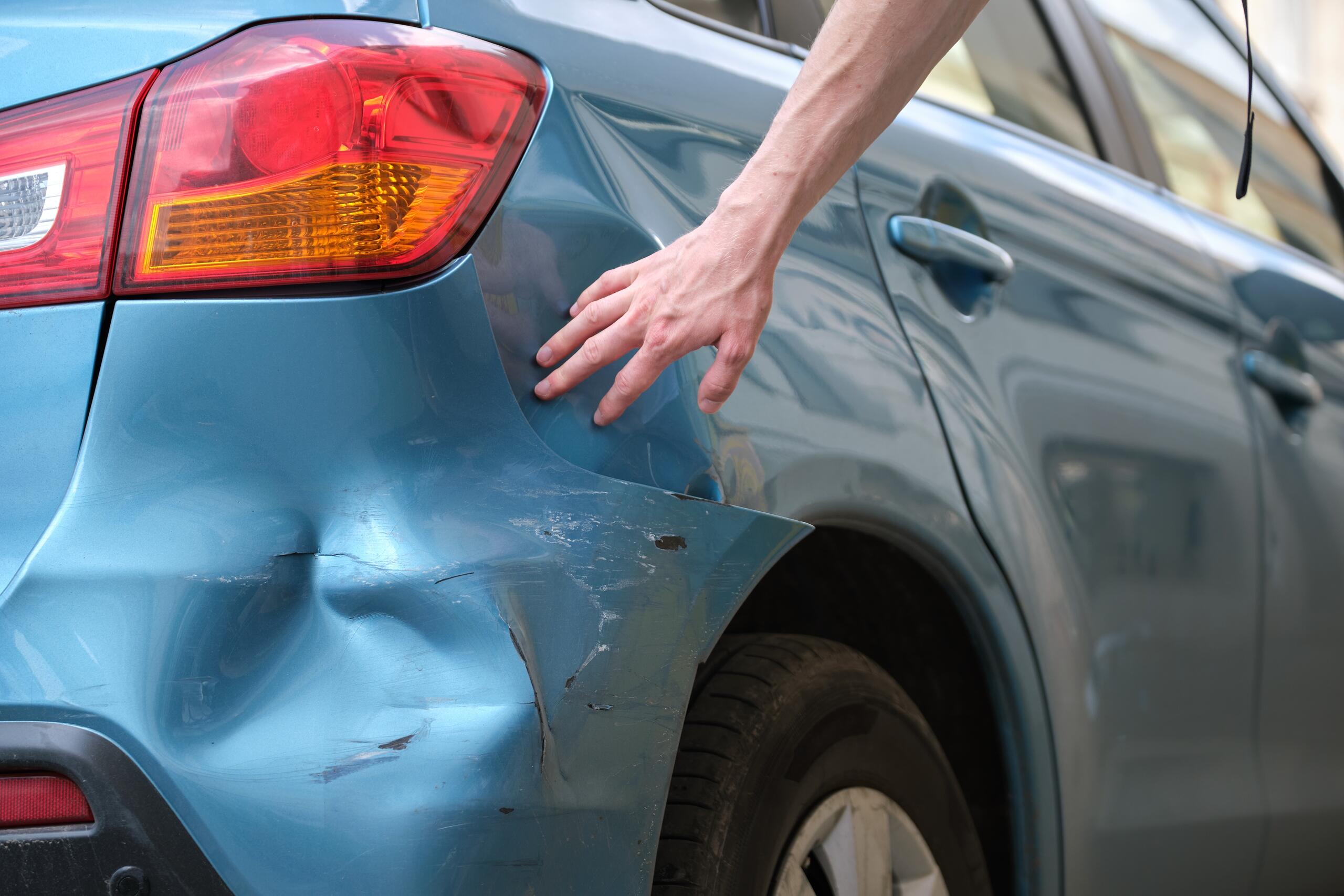 A hand touching a dented blue car bumper, illustrating vehicle damage assessment in the car accident claims process in Missouri.